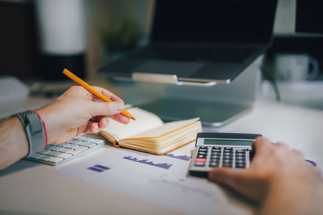 Hands using a calculator and making notes beside a laptop while budgeting for a new website